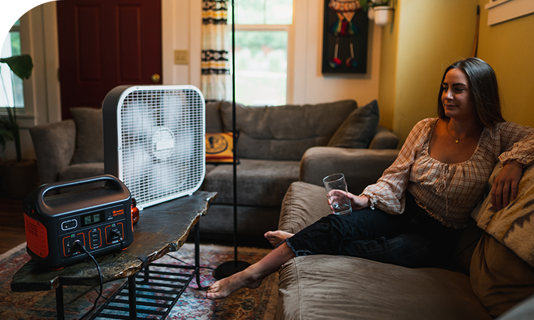 A Jackery Portable Power Station sits on a coffee table, powering a floor fan for a woman relaxing on a couch indoors.