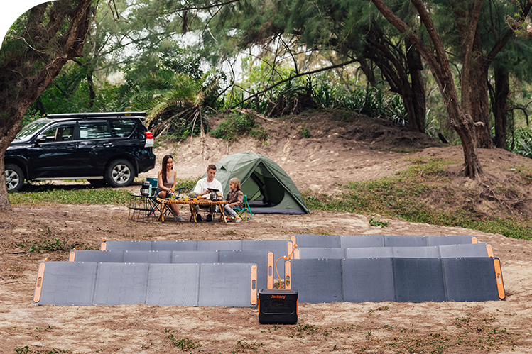 A Jackery Explorer Portable Power Station is set up on the ground connected to multiple solar panels at a forest campsite with a tent, vehicle, and family nearby.