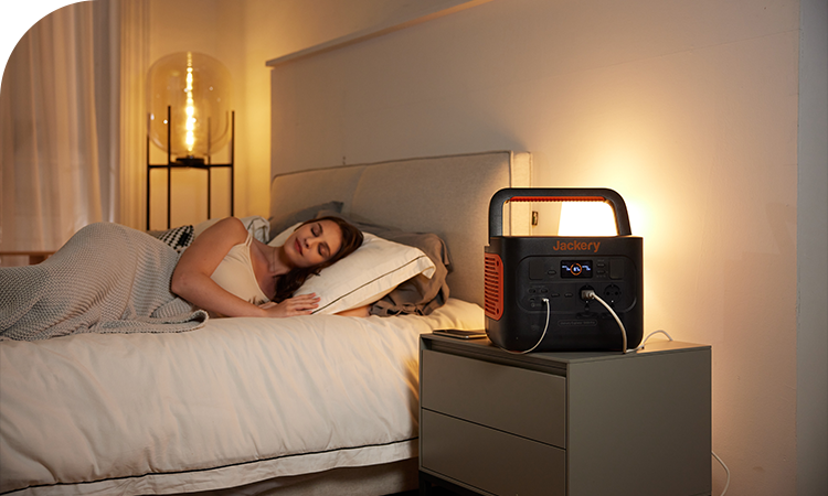 A Jackery Explorer Portable Power Station sits on a nightstand, supplying power to a bedside lamp and a phone while a woman sleeps in bed.