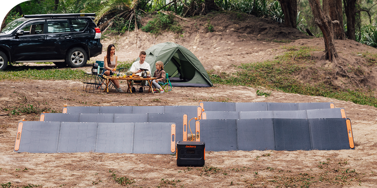 A Jackery Explorer Portable Power Station is set up on the ground connected to multiple solar panels at a forest campsite with a tent, vehicle, and family nearby.