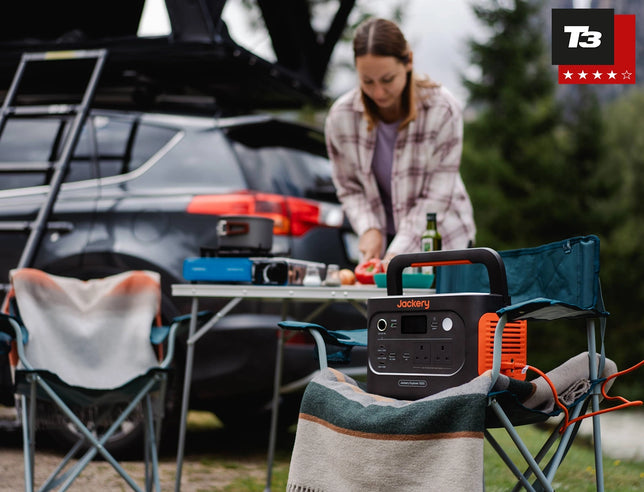 A woman sets up a small outdoor cooking table beside a car in a campsite setting, with a Jackery portable power station placed in the foreground on a chair. The scene includes folding chairs, a blanket, trees, and a T3 rating badge in the top right corner.