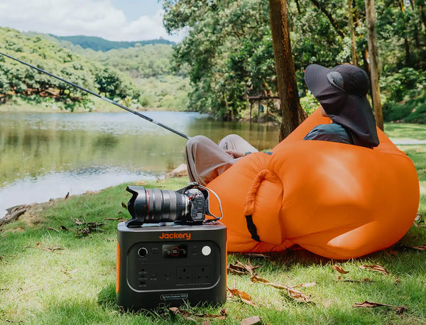 A person relaxes in an orange inflatable lounge chair by a lake, with a Jackery portable power station and camera in the foreground on the grass.