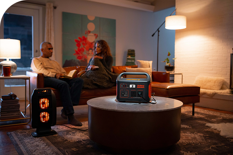 A Jackery Explorer Portable Power Station on a coffee table powers a space heater in a living room while a couple relaxes on a couch.