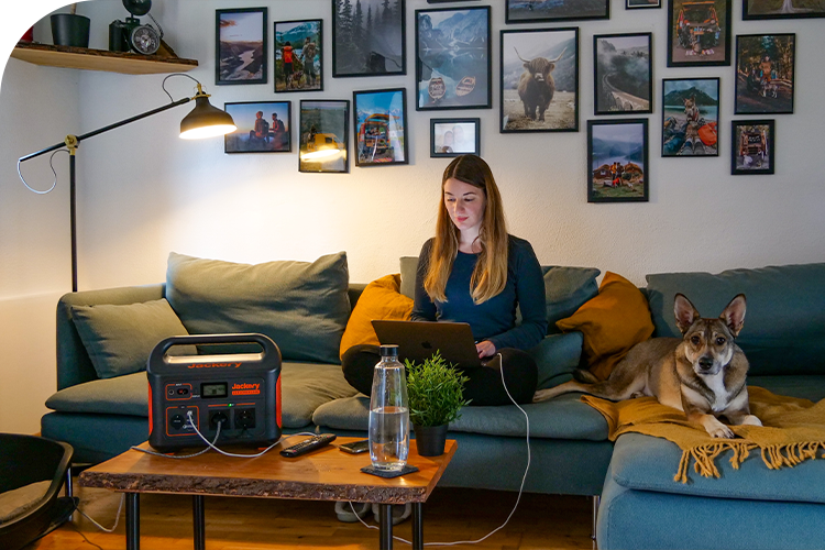 A Jackery Explorer Portable Power Station on a coffee table powers a laptop for a woman and a floor lamp in a living room.
