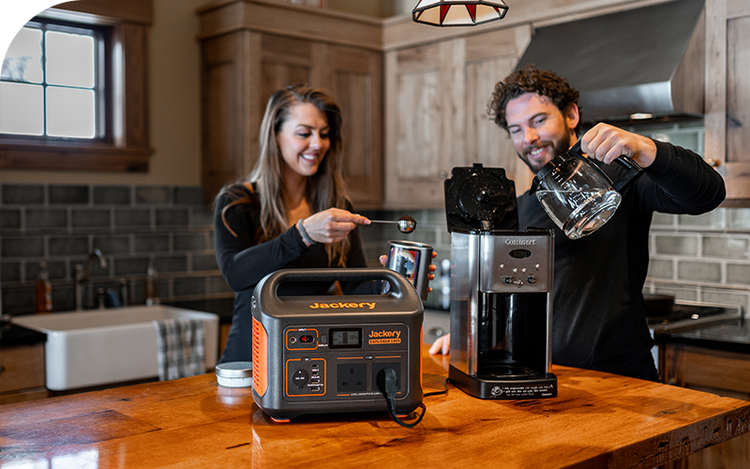 A Jackery Explorer Portable Power Station on a wooden kitchen counter powers a coffee maker for a man and woman preparing coffee at home.