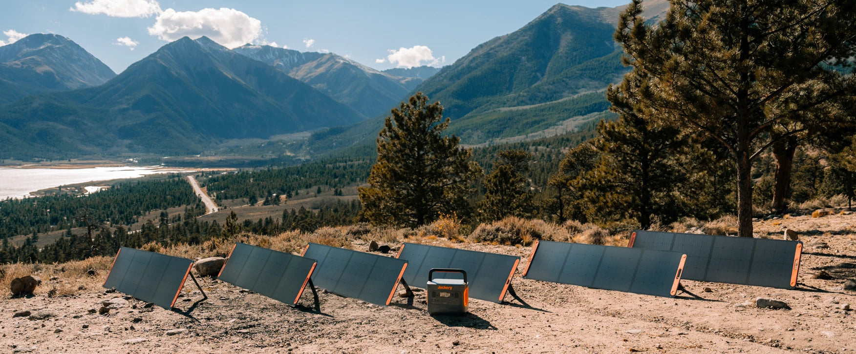 Several Jackery solar panels and a portable power station set up in a scenic outdoor location with mountains and trees in the background.