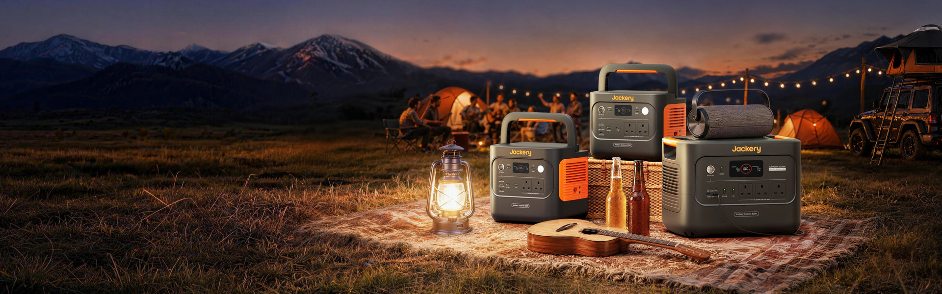 Three Jackery Explorer v2 portable power stations (1000 v2, 2000 v2, and 3000 v2) displayed outdoors on a patterned rug with camping gear, beer bottles, and a guitar, with camping tents, a vehicle with a rooftop tent, and a sunset mountain backdrop in the background.