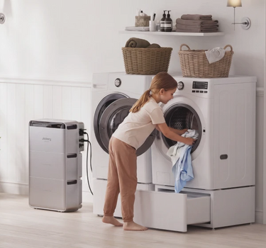A child in beige pajamas stands in a bright laundry room, placing a light blue shirt into the drum of a white front-loading washing machine. A SolarVault 3 Solar Battery stands to the left, and baskets of folded towels and a shelf with toiletries are visible in the background.