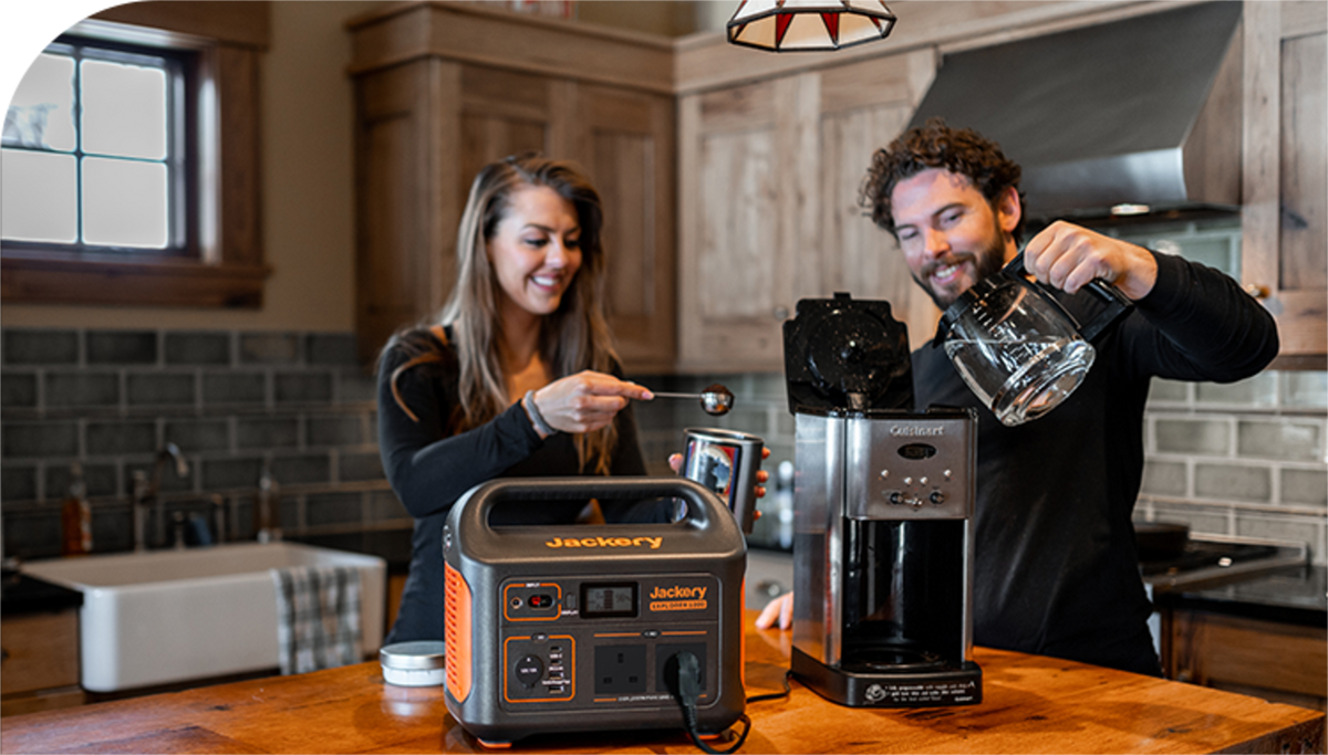 A Jackery Explorer Portable Power Station on a wooden kitchen counter powers a coffee maker for a man and woman preparing coffee at home.