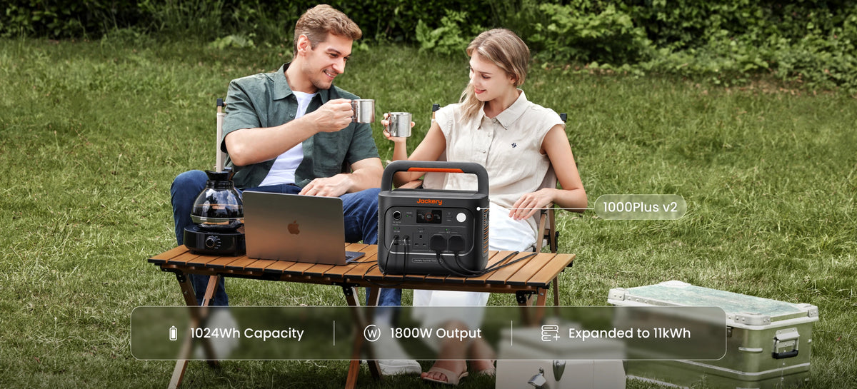 A man and a woman sit outdoors on a grassy lawn around a small table setup,  a Jackery 1000 Plus V2 portable power station is powering for their devices.
