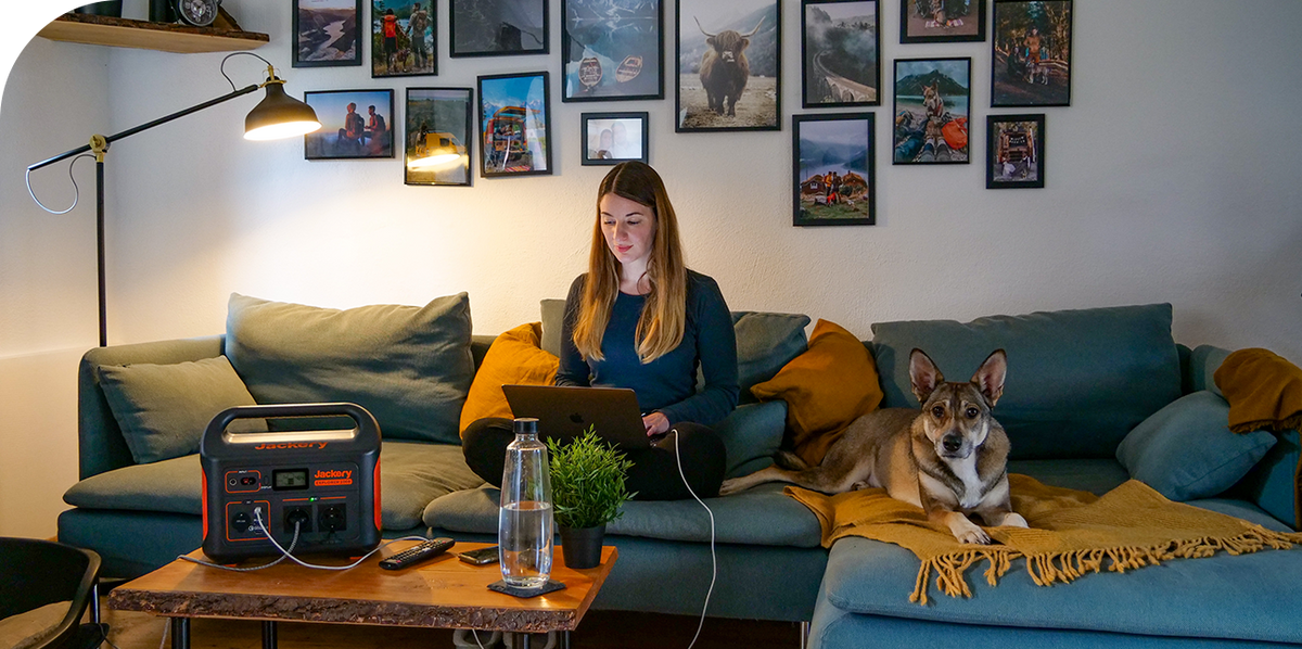 A Jackery Explorer Portable Power Station on a coffee table powers a laptop for a woman and a floor lamp in a living room.