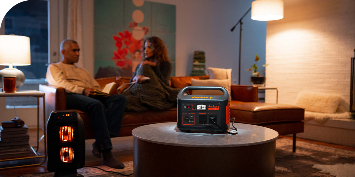 A Jackery Explorer Portable Power Station on a coffee table powers a space heater in a living room while a couple relaxes on a couch.
